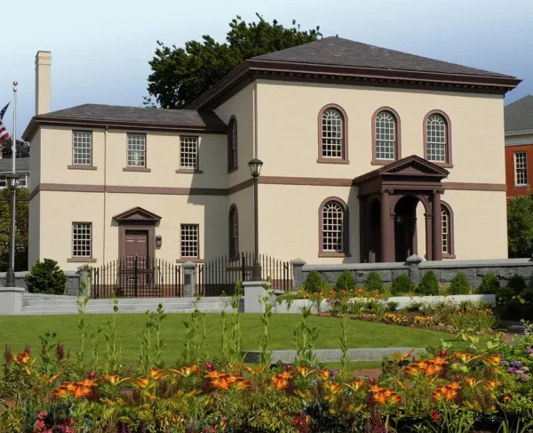 A beige building with brown trim and a columned portico stands behind a garden of bright flowers and neatly trimmed shrubs, bordered by a wrought‑iron fence and stone posts.