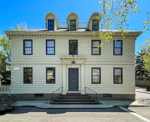 A light‑colored historic building with dark shutters and a central double‑door entrance sits on a corner lot, featuring three dormer windows on the roof and a short staircase leading up from the sidewalk.