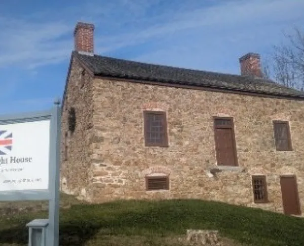 A rectangular stone house with two chimneys stands on a grassy lawn, with a sign in front identifying it as the 1759 Vought House, a Loyalist home from Revolutionary times.