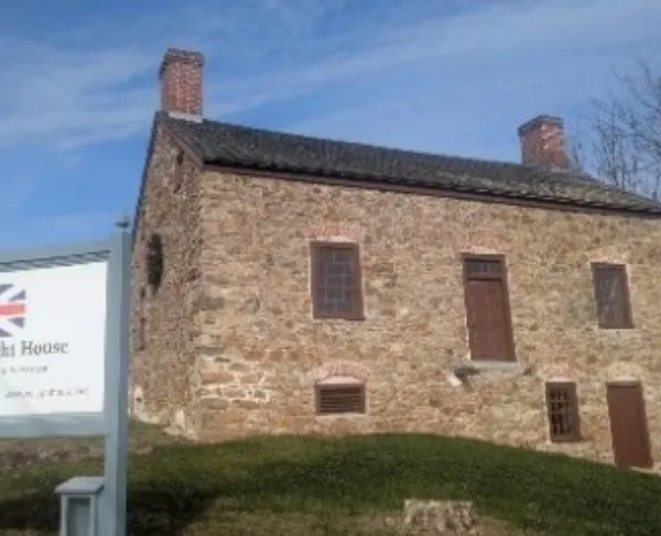 A rectangular stone house with two chimneys stands on a grassy lawn, with a sign in front identifying it as the 1759 Vought House, a Loyalist home from Revolutionary times.