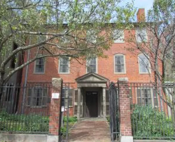 A three‑story, two chimney brick building stands behind a black metal fence and brick pillars, partly shaded by leafy trees.