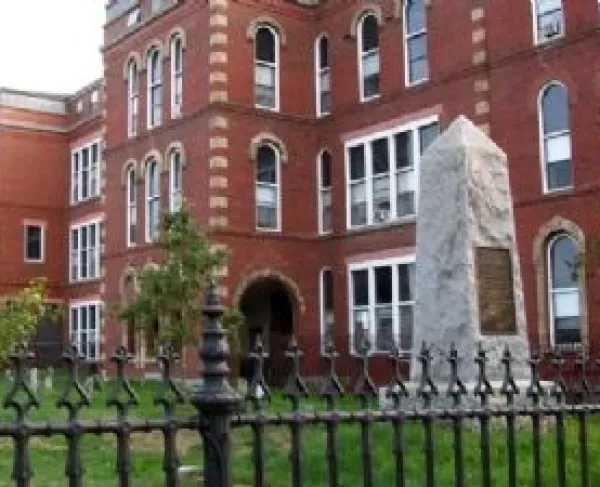 A tall red‑brick historic building with arched windows stands behind a small stone monument shaped like an obelisk. The monument has a metal plaque on its front and is enclosed by a black wrought‑iron fence.