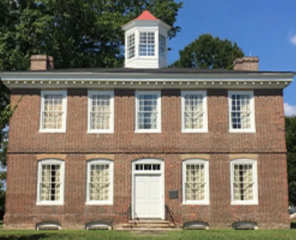 A two‑story brick Federal style building with white‑trimmed windows and a central cupola stands on a grassy lawn beneath a bright blue sky.