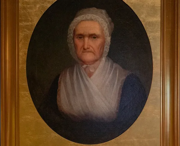 A painting of Charlotte Robertson, an older woman in a dark dress with a white lace kerchief and cap. The painting is in a gold frame. 