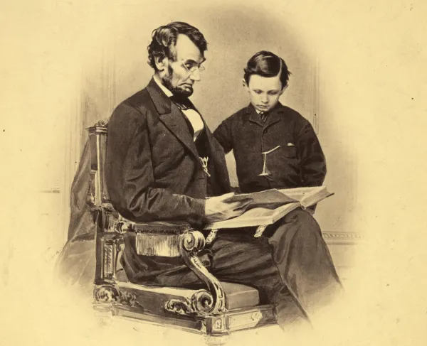 President Abraham Lincoln sits wearing spectacles beside his young son Tad, examining a book altered in the photograph to resemble a Bible.