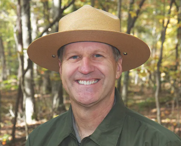 Smiling man wearing a broad-brimmed National Park Service ranger hat and green uniform, outdoors in a wooded setting.