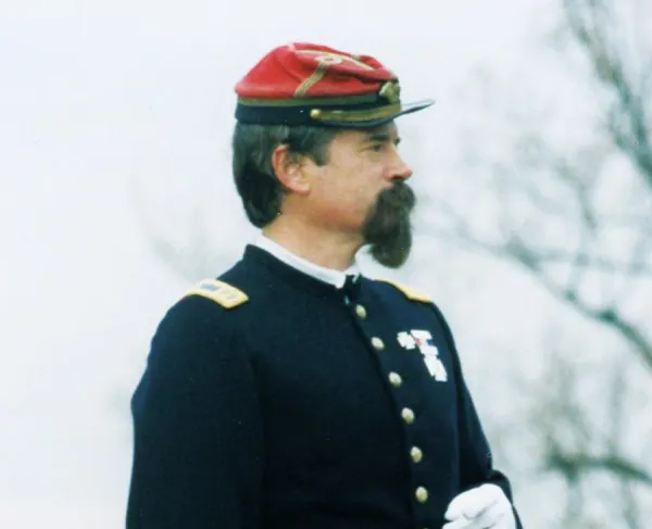 Man with dark hair and a mustache wearing a Civil War-era military uniform and red kepi-style cap.