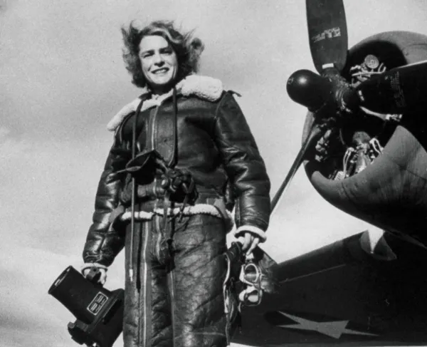 Black and white photo showing a woman in a pilot's jacket with a camera, standing in front of airplane propeller