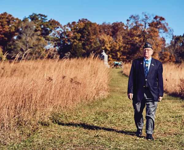 World War II Veteran in a blazer and hat walks a field with a monument in the background