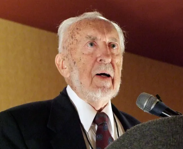 Man with white hair and a white beard speaking at a podium into a microphone, wearing a dark suit, white shirt, and patterned tie.