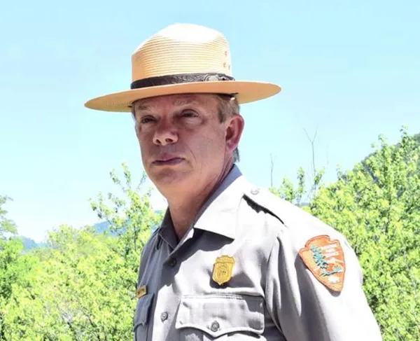 Man wearing a National Park Service uniform and broad-brimmed ranger hat, outdoors against trees and a blue sky.