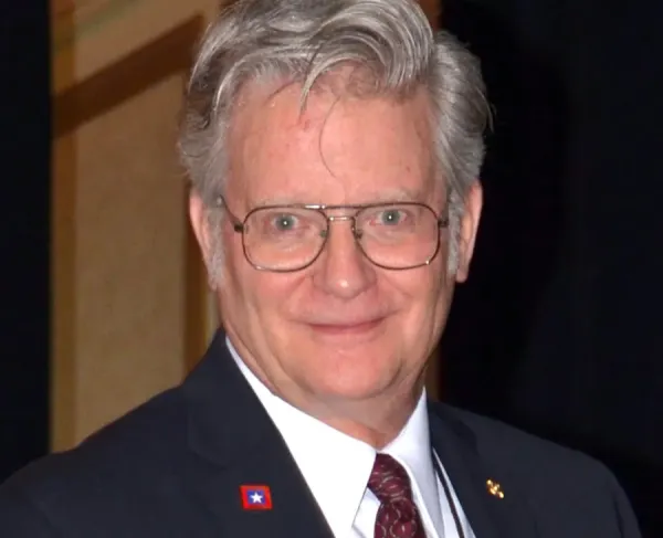 Man with gray hair and glasses smiling at the camera, wearing a dark suit, white shirt, and patterned tie.