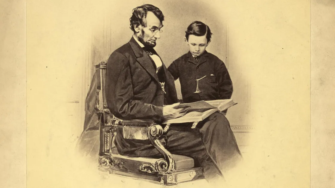 President Abraham Lincoln sits wearing spectacles beside his young son Tad, examining a book altered in the photograph to resemble a Bible.