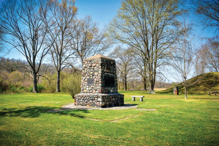 Short obelisk monument covered in rocks in a field with trees and mound seen in the distance