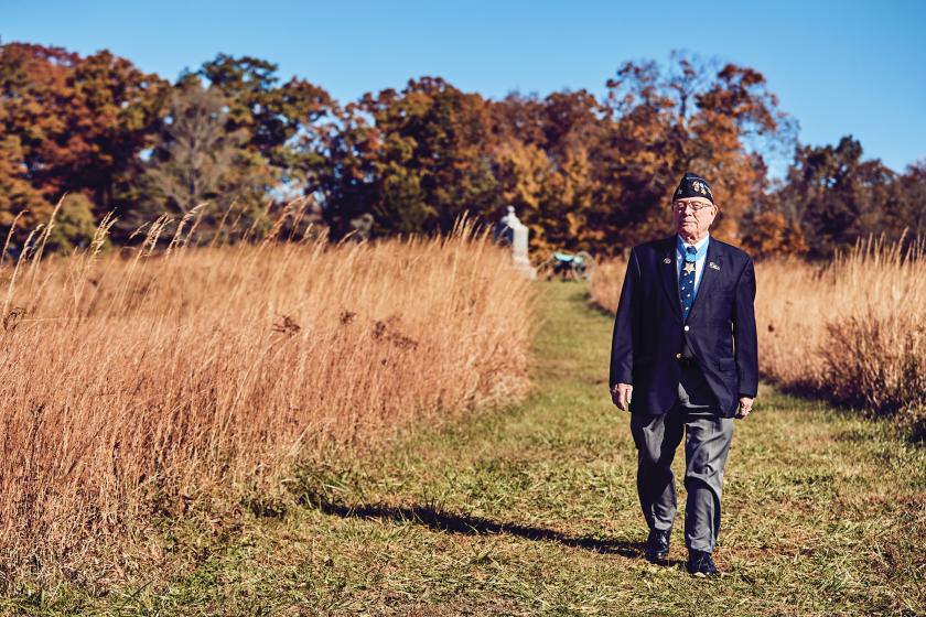 World War II Veteran in a blazer and hat walks a field with a monument in the background
