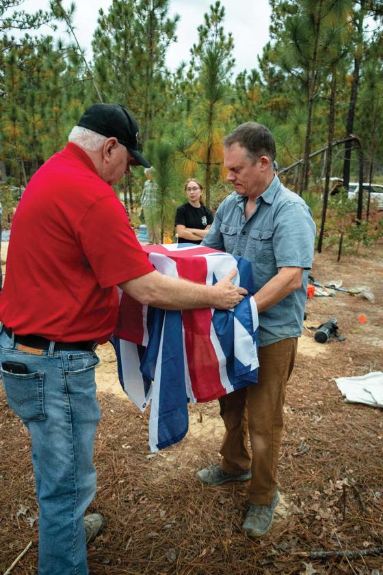 SCBPT staff member and military veteran Rick Wise (LEFT) drapes the remains of a British loyalist with the Kings Colors while Dr. Bill Stevens, deputy coroner, Richland County prepares to carry the remains to the coroner’s van.