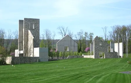 A cluster of modern stone and concrete buildings sits on a gently sloping green lawn. The structures have clean geometric lines, including tall rectangular towers and gabled‑roof sections. 