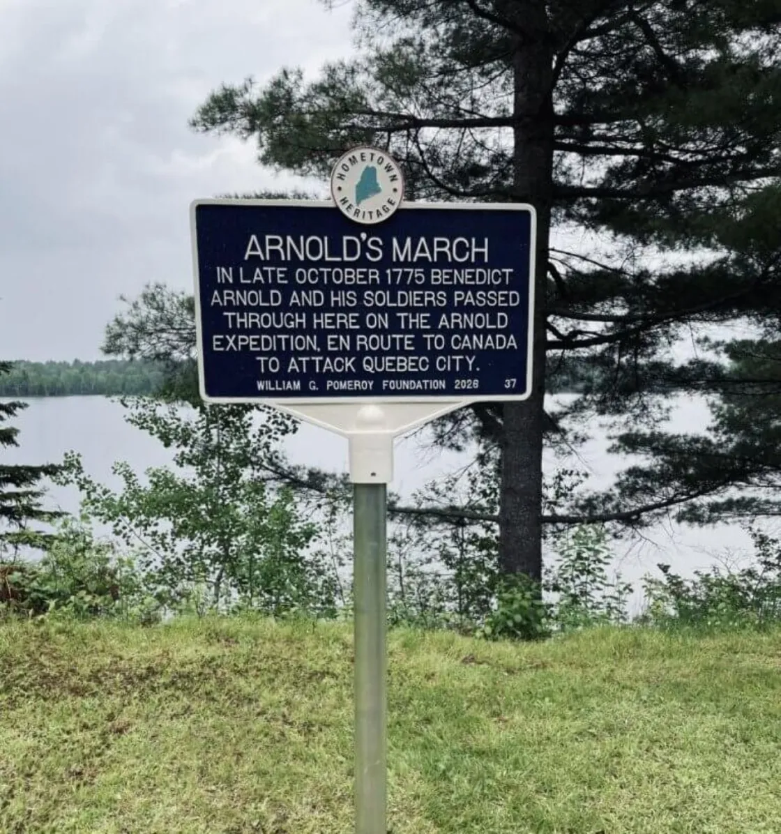 A blue “Arnold’s March” historical marker stands on a grassy lakeside clearing, surrounded by trees under a clear sky.