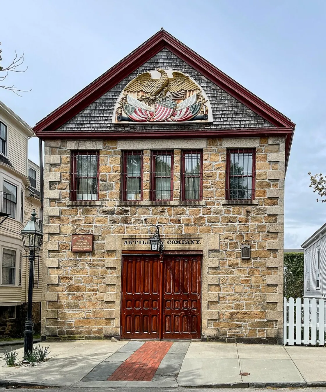 A small historic stone armory built of rough gray blocks with red trim along the gabled roof. Centered near the roof is a large carved and painted patriotic emblem featuring an eagle with outstretched wings and crossed American flags.
