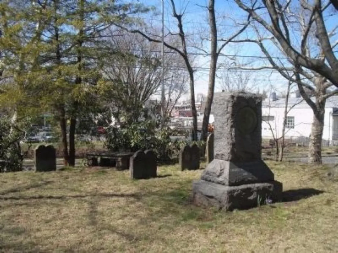 A large stone monument with an engraved circular emblem stands among rows of headstones in a small grassy cemetery bordered by leafless trees and nearby buildings.