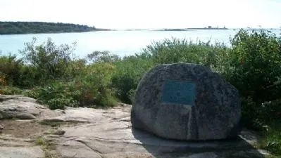 A large rock with a metal plaque sits on a rocky shoreline overlooking calm water, surrounded by low greenery and distant tree‑covered land.