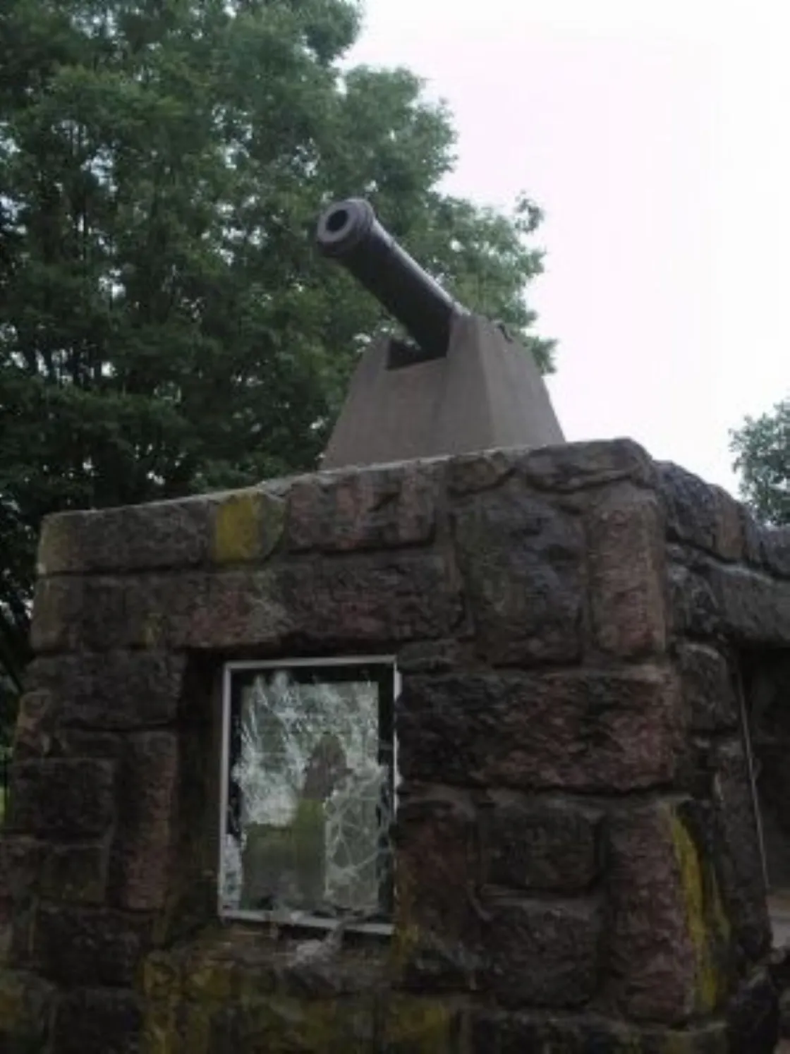 A black cannon sits atop a small stone structure with a glass window, surrounded by green trees under an overcast sky.