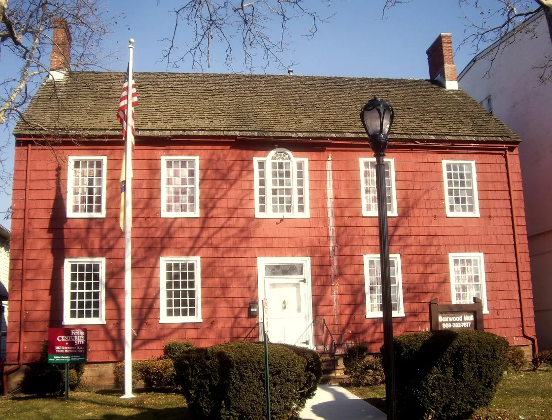 A red historic house with white trim and twin brick chimneys stands behind a flagpole and walkway lined with bushes.