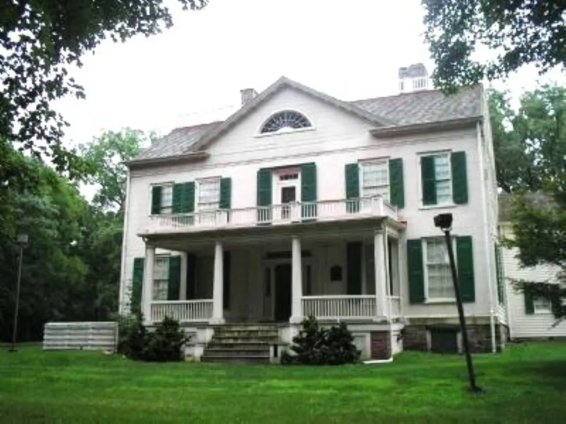 A large white historic house with green shutters, a columned front porch, and a balcony stands on a grassy lawn surrounded by trees.