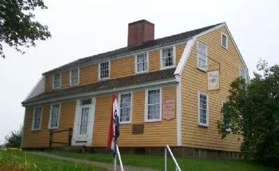 A two‑story yellow wooden house with white trim stands behind a small walkway. The building has a steep roof, a central brick chimney, and evenly spaced white‑framed windows.