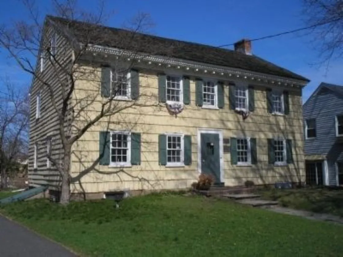 A yellow colonial‑style house with green shutters and a steep dark roof stands behind a leafless tree, with a stone walkway leading to its green front door.