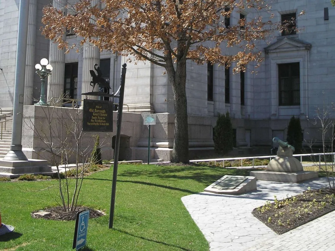 A light‑stone courthouse stands behind a grassy area with a tree, a small cannon monument, and a historical marker noting the site of an early borough courthouse built before the Revolution.