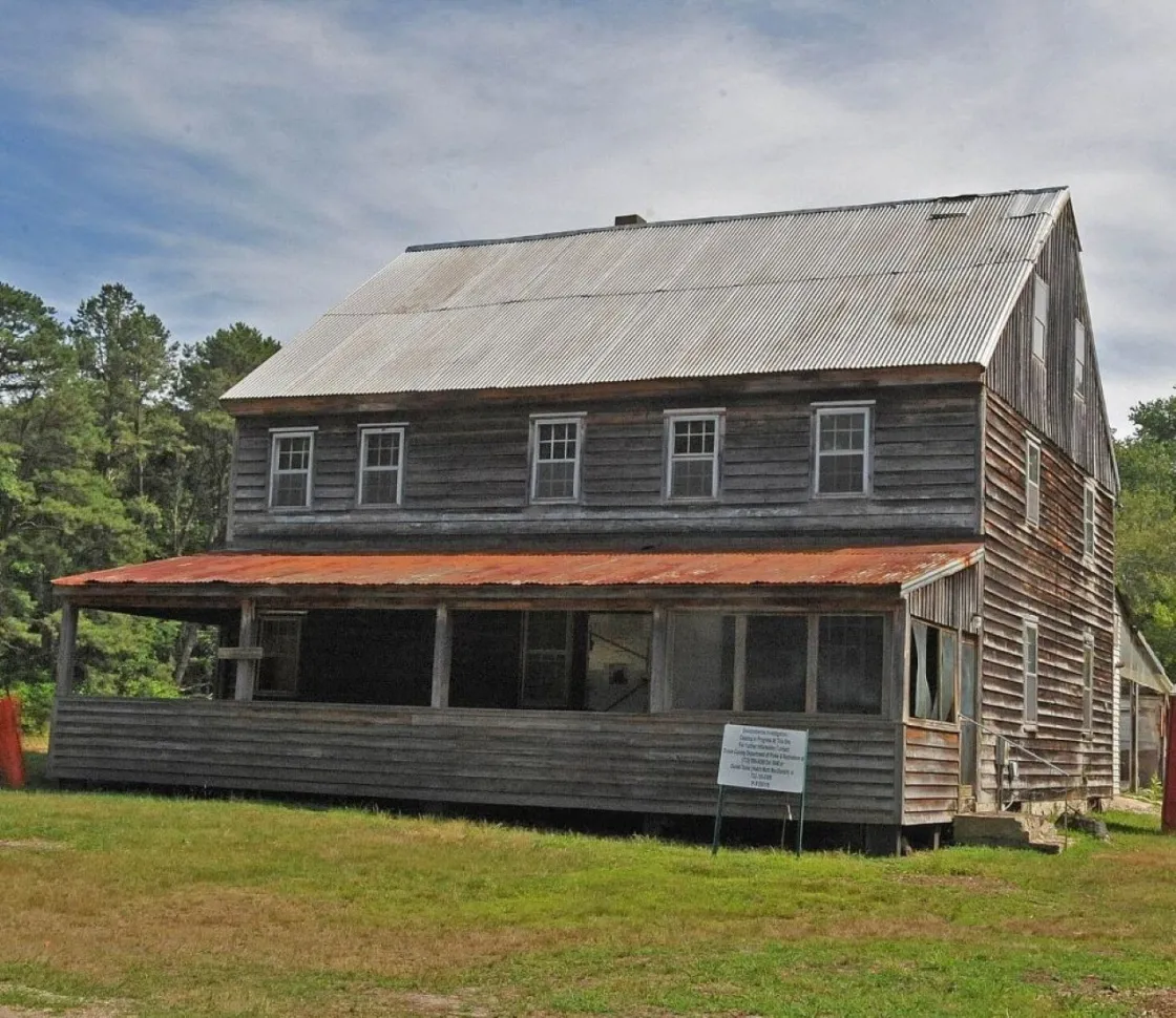 An old two‑story wooden building with a rusted metal porch roof stands in a grassy clearing.