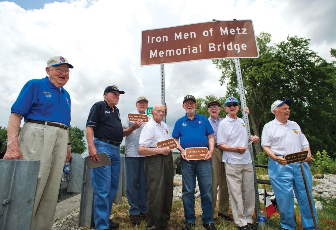 A group of eight veterans stands with a sign that reads Iron Men of Metz Memorial Bridge