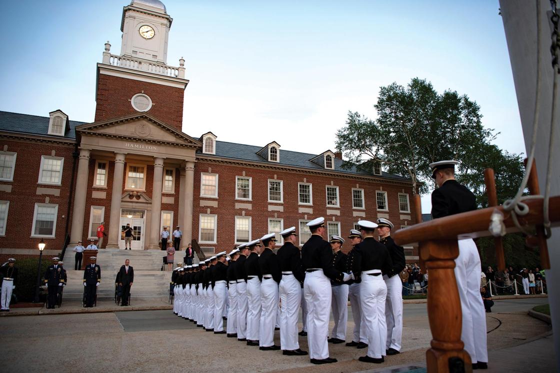 Two rows of cadets in uniform face each other in front of a building with a tower