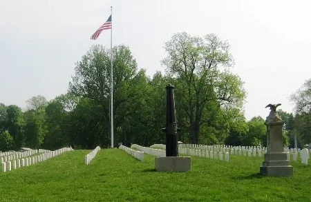 Rows of white headstones line a green cemetery lawn, arranged in straight, even rows. A tall flagpole flying the United States flag stands in the center background. In the foreground, a vertical cannon monument and a stone marker topped with an eagle sculpture sit among the grave.