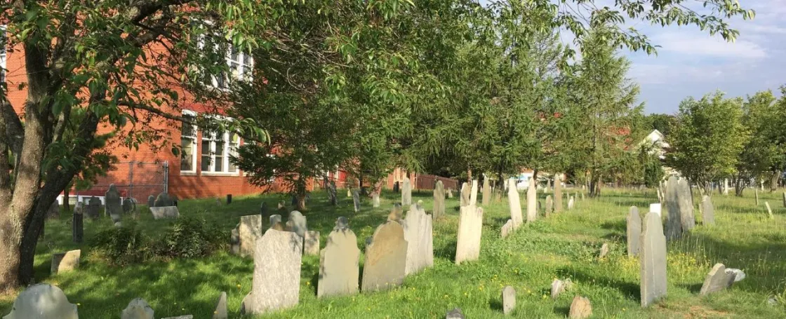 Historic cemetery with rows of weathered gravestones standing in a grassy, tree-shaded burial ground beside a large red-brick building, illuminated by late afternoon sunlight.