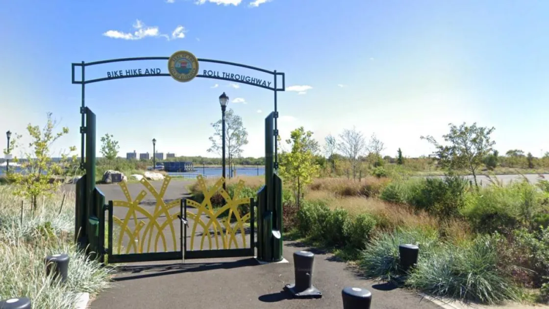 A decorative metal gateway features tall yellow metal shapes resembling stylized reeds, with a circular emblem above of the Seal of Elizabeth.  Beyond the gate, the path leads toward a waterfront lined with trees, shrubs, lampposts, and distant buildings under a bright blue sky.