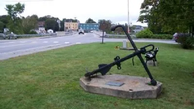A large black metal anchor sits upright on a square concrete base in a small grassy roadside park. A bronze plaque is mounted on the front of the base, and a wooden bench stands nearby. 