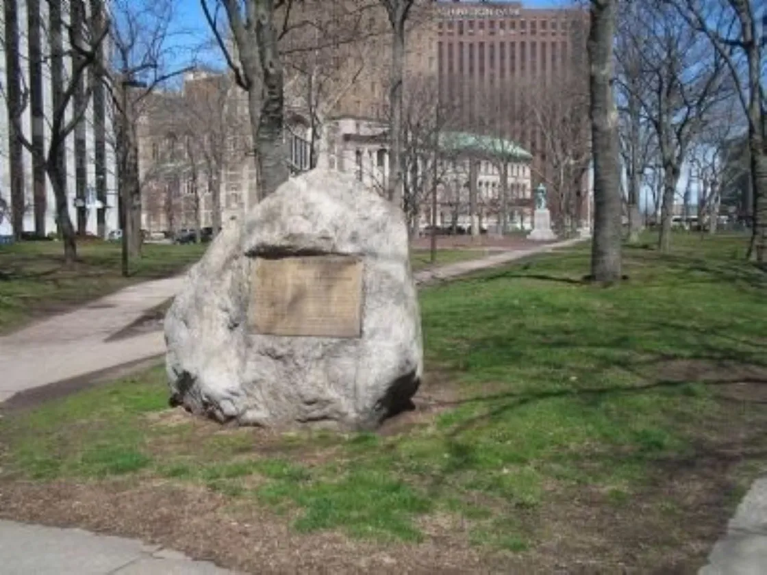 A large boulder with a metal plaque stands in a grassy urban park surrounded by trees and tall buildings.