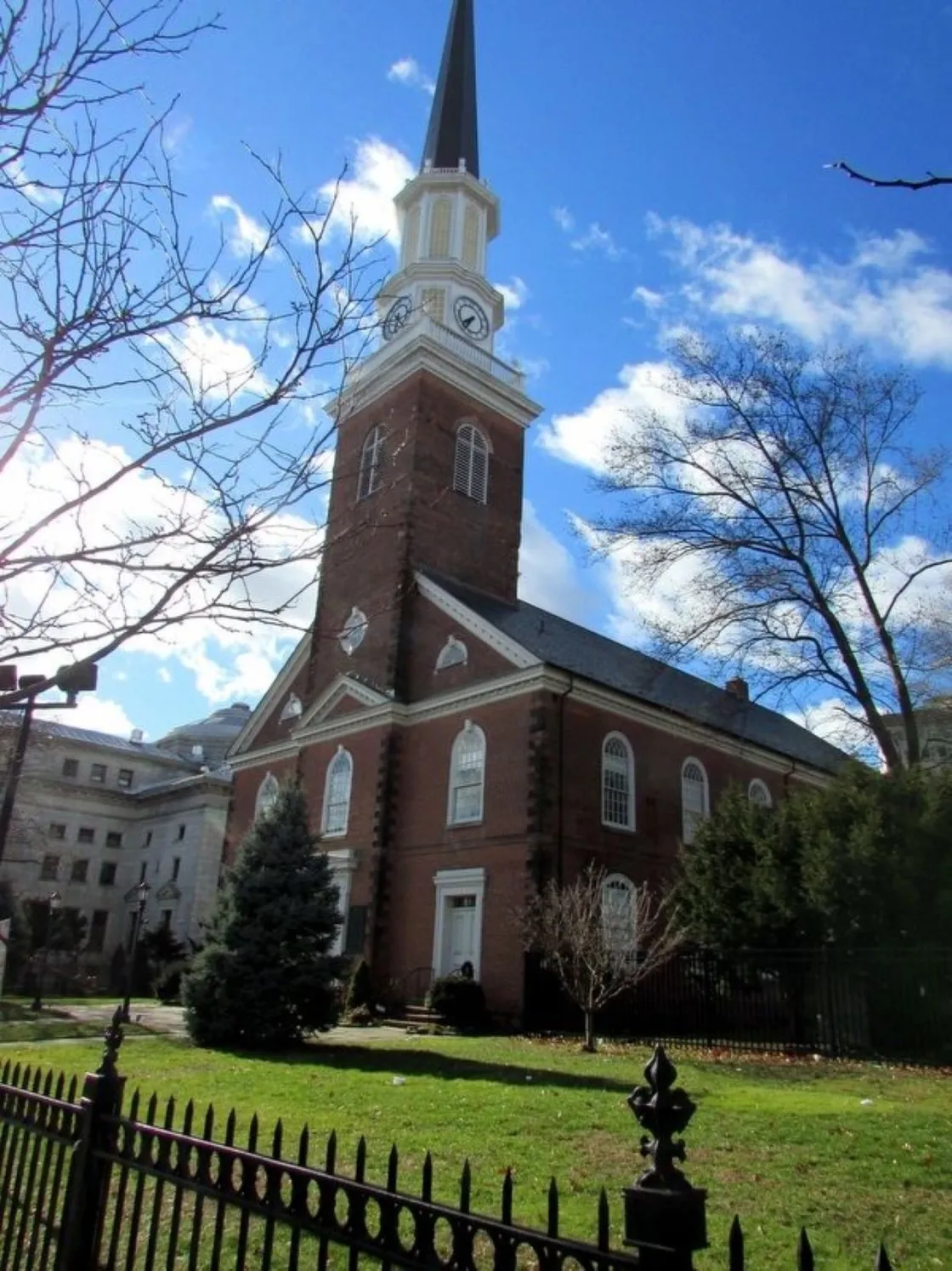 A tall brick church with a white clock‑tower steeple rises above a fenced grassy area with trees under a bright blue sky.