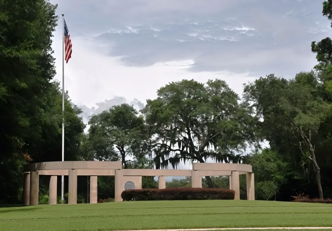 A circular memorial made of tall stone columns with a ring-shaped structure resting on top of the columns. A manicured lawn and trimmed hedge sit in front, and a flagpole flying the United States on the left. 