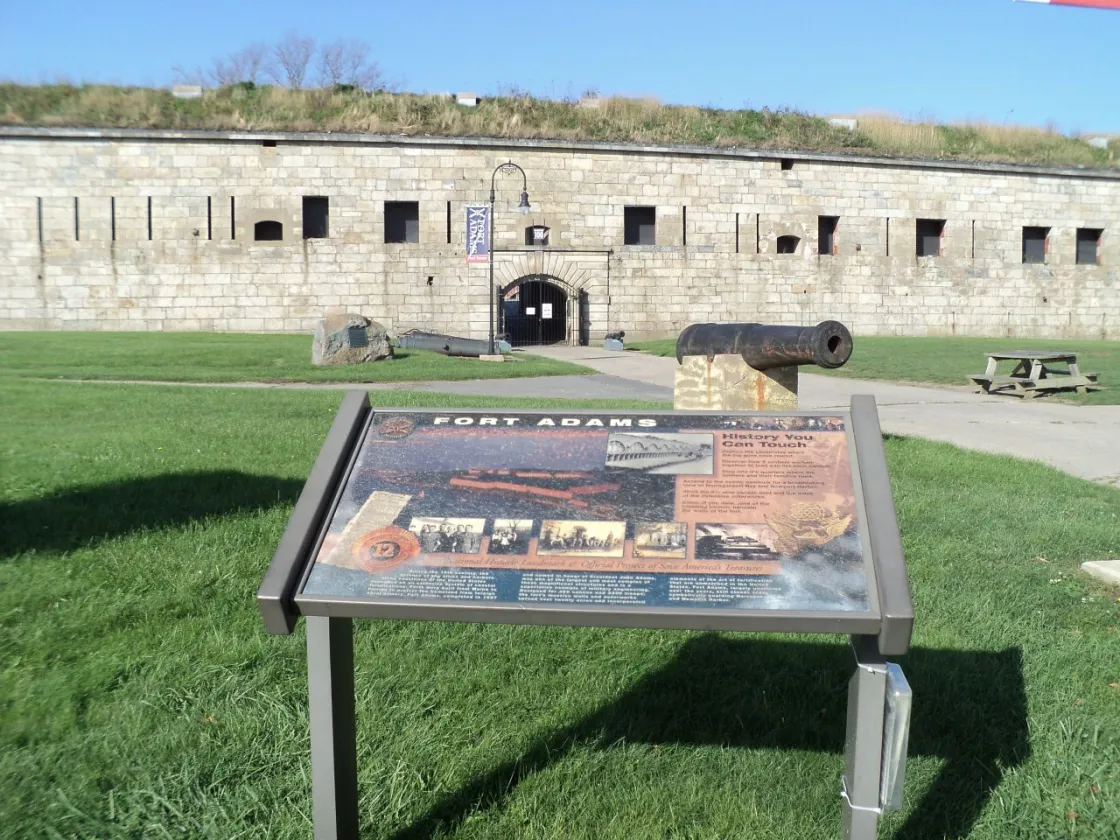 A stone fort wall with an arched entrance rises behind a grassy area where an informational sign, an old cannon, and a picnic table sit under a bright blue sky.