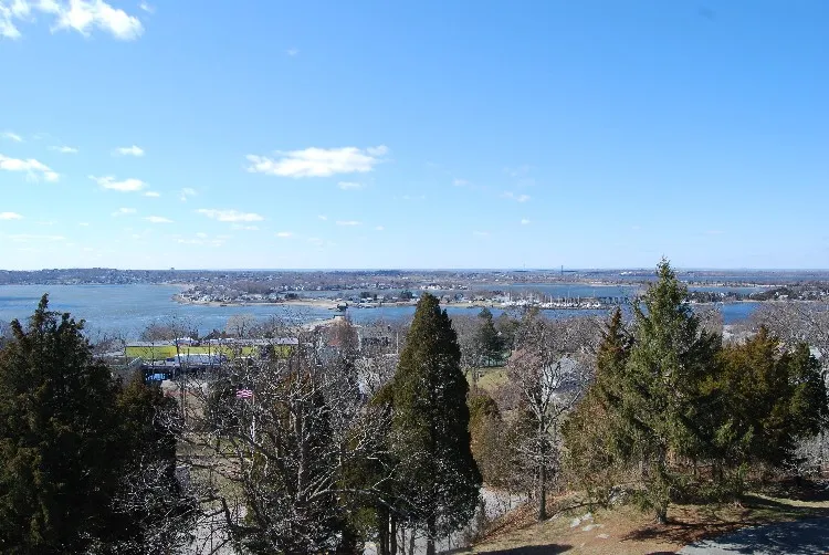 A wide view of a calm blue bay stretches toward distant shoreline homes, framed in the foreground by tall evergreens and leafy trees beneath a sky dotted with white clouds.