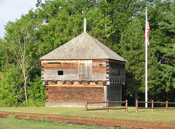 A two‑story wooden blockhouse stands beside a flagpole on a grassy area near dense trees in the background.