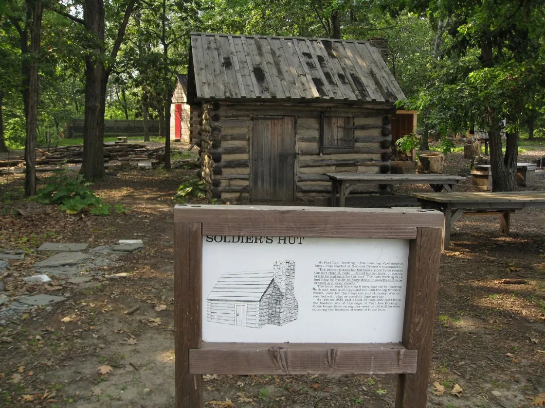A small reconstructed Revolutionary War–era log cabin stands in a wooded clearing. The structure is labeled “Soldier’s Hut” on an informational sign in front of it. 