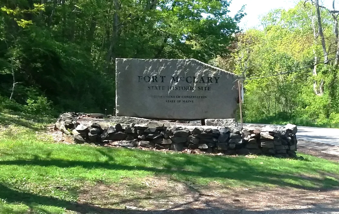  A stone sign reading “Fort McCleary State Historic Site” stands beside a road, surrounded by a low stone wall, grass, and trees.