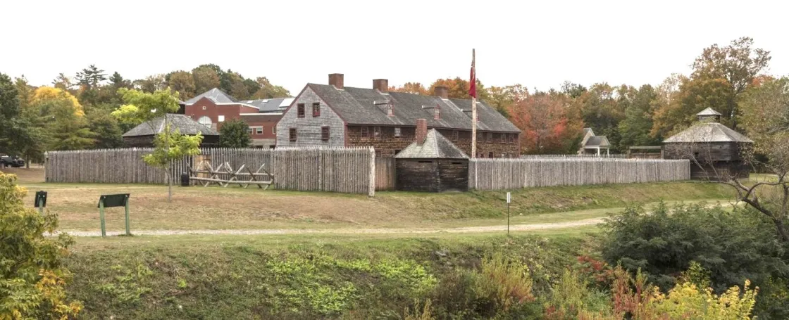 A colonial-era fort enclosed by a tall wooden palisade stands amid autumn-colored trees, with weathered timber buildings, a flagpole, and grassy grounds viewed from across a shallow ditch.