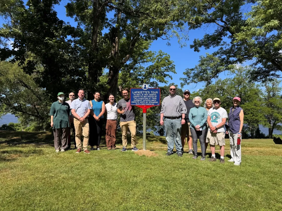 A blue historical marker outlined in red with white lettering titled “Lafayette’s Tour” stands on a lawn in front of a tree surrounded by people commemorating Lafayette's visit to this city.