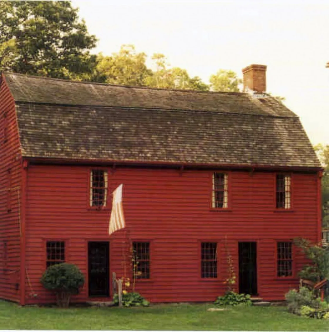 A red wooden colonial‑style house with a steep shingled roof and small‑paned windows stands amid green grass and trees, with an American flag mounted near one of its front doors.