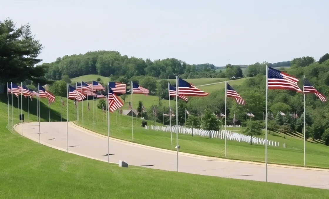 A roadway curves through a military cemetery, lined with tall flagpoles carrying American flags, with rows of white headstones stretching across the green hillside.